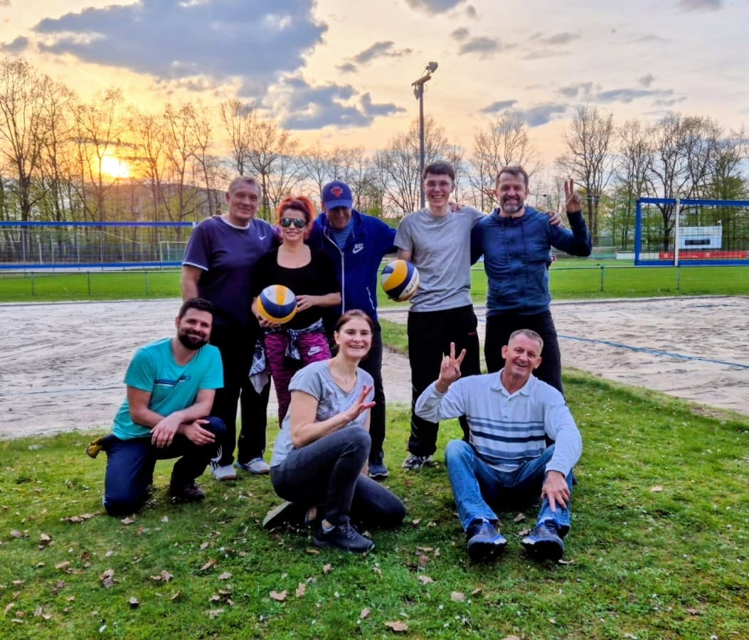 Group of eight adults on a grassy field at a volleyball court posing with two yellow-blue volleyballs at sunset.