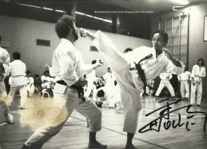 Black-and-white dojo scene: two karate practitioners in white gi sparring as a high kick flies toward the opponent; classmates watch in the background.