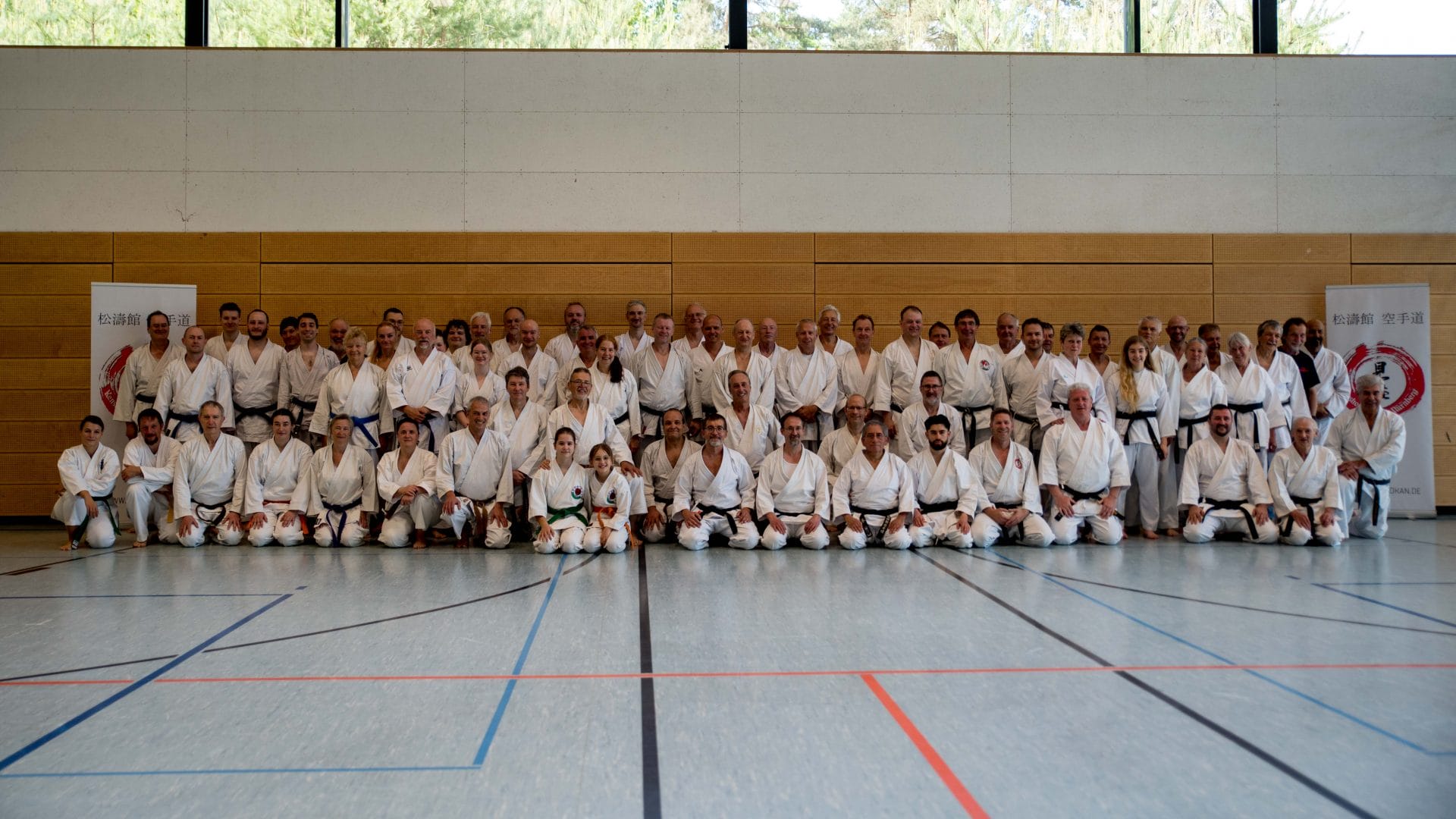 Group photo of a large martial arts class: dozens of students and instructors in white gis lined up in a gym, posing for the picture.